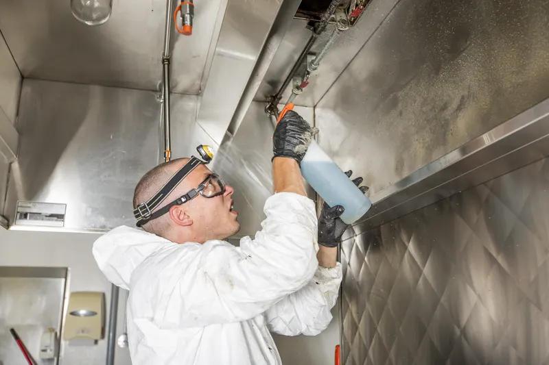 Technician in a headlamp and white coveralls spraying degreaser up under the edge of a stainless kitchen exhaust canopy