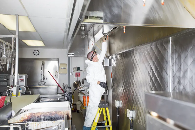 Hood cleaning technician on a step ladder cleaning a stainless canopy in a full commercial kitchen
