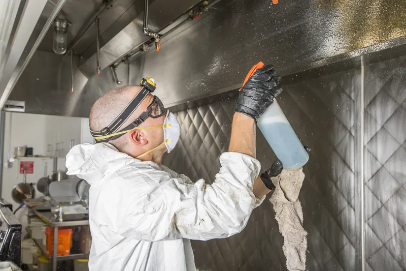 Technician spraying and wiping the interior of a stainless exhaust hood with commercial cooking equipment behind him